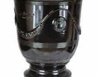 May include: A dark brown, glossy ceramic planter with a wide, flared rim and a pedestal base. The exterior features a raised, decorative relief pattern consisting of draped swags and circular medallions encircling the upper body of the vessel.
