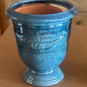 May include: A blue glazed ceramic planter with a wide rim and a pedestal base. The exterior features a relief design of wheat stalks and the handwritten text "la Madeleine" and "fudure" in black script. The interior shows a natural terracotta finish.