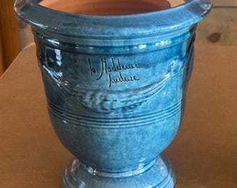May include: A blue glazed ceramic planter with a wide rim and a pedestal base. The exterior features a relief design of wheat stalks and the handwritten text "la Madeleine" and "fudure" in black script. The interior shows a natural terracotta finish.