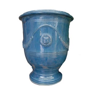 May include: A glossy blue ceramic planter with a wide, flared rim and a tapered base. The exterior features a raised, arched decorative molding and a central circular emblem with the embossed text POTERIE LA MADELEINE ANDUZE GARD.