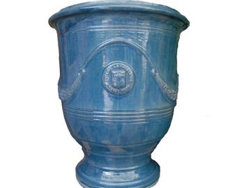 May include: A glossy blue ceramic planter with a wide, flared rim and a tapered base. The exterior features a raised, arched decorative molding and a central circular emblem with the embossed text POTERIE LA MADELEINE ANDUZE GARD.
