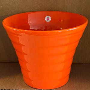 May include: A bright orange ceramic planter with a glossy finish and a ribbed, tapered design. The interior features a small circular white sticker with the text Pottery Company Los Angeles printed in black and a small red graphic in the center.