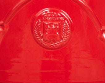 May include: A bright red ceramic surface featuring a circular embossed emblem in the center. The emblem displays the text LA MADELEINE surrounded by a laurel wreath design. The object has a smooth, glossy finish with curved, raised lines.