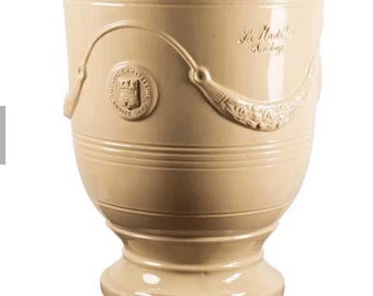May include: A cream-colored ceramic planter with a flared rim and a pedestal base. The exterior features a raised relief design of draped garlands and a circular emblem with the text La Madeleine and a crest with three towers and a shield.