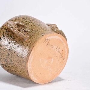 May include: A handcrafted, glazed stoneware pottery jug with a textured surface and two handles. The jug is primarily olive green with brown accents. The bottom of the jug is unglazed and features the artist's signature.