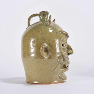 May include: A handcrafted, green ceramic jug with a devilish face design. The jug features a handle, horns, a prominent nose, and a detailed face. The stoneware has a speckled texture and a rustic, antique aesthetic.