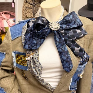 May include: A tan plaid blazer featuring denim patches, a large bow made from blue paisley and checkered neckties, a white pearl necklace, and a gold-tone brooch with pearls. A patch with the text LIMITED EDITION is sewn onto the denim.