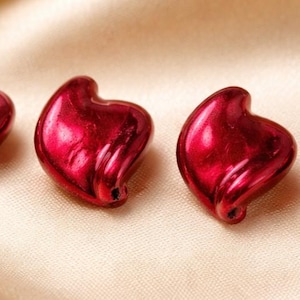 May include: Four individual heart-shaped beads with a glossy, metallic red finish. Each bead features a smooth, rounded surface and a small hole at the bottom point, designed for stringing. The beads are arranged in a horizontal row.