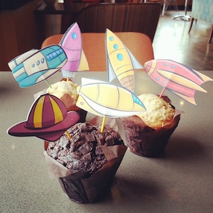 May include: Four cupcakes decorated with colorful paper rockets and a fire hat. The cupcakes are on a light gray surface.