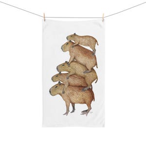 May include: A white towel featuring a stack of five capybaras, each one atop the other. The capybaras are depicted in shades of brown and tan, with detailed fur textures. The towel is hanging from a clothesline.