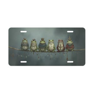 May include: A rectangular license plate featuring a digital illustration of six frogs perched on a thin branch. The frogs are adorned with various crowns and ornate clothing, with delicate hanging chains and charms suspended from the branch below.