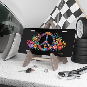 May include: Black license plate featuring a colorful peace sign design surrounded by flowers. The peace sign is filled with a floral pattern. The flowers are in shades of pink, yellow, blue, and orange. The plate is displayed on a wooden stand.