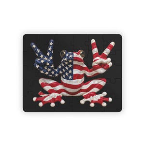 May include: A rectangular jigsaw puzzle featuring a frog with its skin patterned like the American flag, including red and white stripes and white stars on a blue field. The frog is positioned centrally, holding both front limbs up in peace signs.
