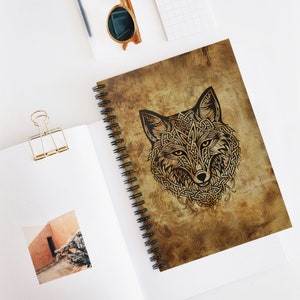 May include: A spiral-bound notebook with a detailed illustration of a fox's head in a Celtic knotwork design. The cover has a distressed, aged paper appearance with a brown and tan colour palette. The notebook is open, revealing blank pages.