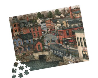 May include: A jigsaw puzzle depicting a town scene with red brick buildings, a blue house, a stone bridge over a river, and cars on a winding road. Several loose puzzle pieces are scattered at the bottom left corner of the rectangular image.