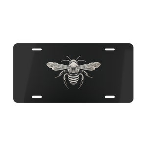 May include: Black and white illustration of a bee on a black license plate. The bee is in the center of the plate and has its wings spread out.
