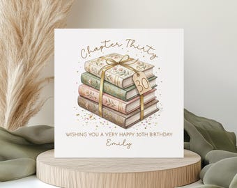 Puede incluir: Tarjeta de cumpleaños cuadrada con una pila de libros atados con una cinta dorada y una etiqueta con el número 30. La tarjeta dice "Capítulo Treinta" y "Deseándote un muy feliz 30 cumpleaños Emily."
