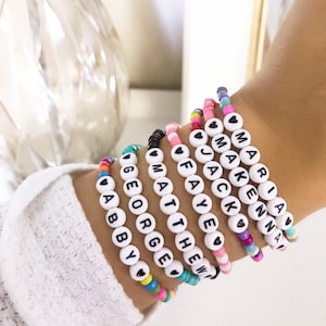 Op de afbeelding: Een set kleurrijke kralenarmbanden met witte kralen die namen spellen, waaronder "Abby", "George", "Matthew", "Fay", "Jack", "Makenna" en "Mariana".