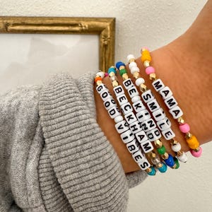 May include: A set of colorful beaded bracelets with different words spelled out on them, including "Mama", "Smile", "Be Kind", "Beckham", "Good Vibes".
