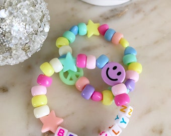 May include: Two colorful beaded bracelets. One bracelet features a smiley face bead, while the other spells out "BROOKLYN" with letter beads. The bracelets include star and peace sign charms and are made with pastel-colored beads.