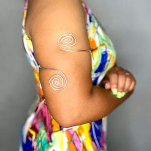 May include: Silver arm cuff featuring two spiral designs. The cuff is worn on the upper arm and forearm. The arm is in front of a colorful patterned dress.