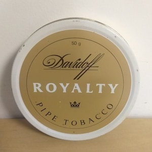 May include: A white round tin with a gold lid. The lid has the text "Davidoff" in a cursive font, followed by the word "ROYALTY" in a bold, sans-serif font. Below the word "ROYALTY" is a crown and the text "PIPE TOBACCO" in a circular pattern.