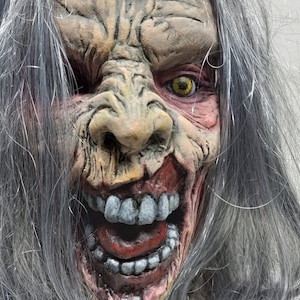 May include: A close-up of a grotesque Halloween mask with a decaying face. The mask features a wrinkled, weathered complexion, exposed teeth, and yellow eyes. Long, gray hair frames the face, adding to the eerie effect.