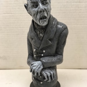 May include: A gray and white bust sculpture of a man with a wrinkled face and a coat. The sculpture is on a black base.