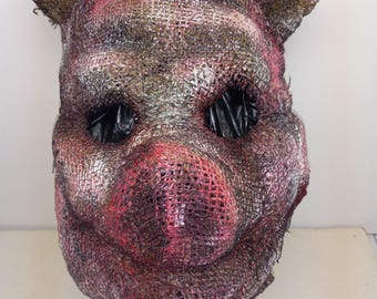 Puede incluir: Una máscara de cabeza completa con forma de cerdo, construida con un material de malla tejida gruesa. La superficie presenta un acabado desgastado y tonal con tonos rojo oscuro, marrón y plata metálica. Tiene dos grandes aberturas huecas para los ojos.