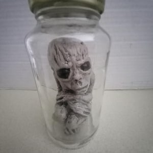 May include: A small, white, humanoid figure with a detailed face is sealed inside a clear glass jar with a gold lid. The lid has the text "After Opening" printed on it.