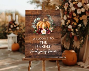 May include: A wooden welcome sign on a wooden easel. The sign features a pumpkin and floral wreath design. The text reads "WELCOME TO THE JENKINS' Thanksgiving Dinner". The sign is set outdoors with fall decorations.