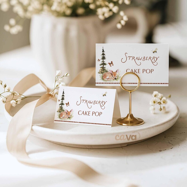 K&ouml;nnte beinhalten: Zwei wei&szlig;e Tischkarten mit dem Text "Strawberry Cake Pop" und einer Aquarellillustration eines Rehs, Schmetterlingen und eines Baumes. Ein goldfarbener Metall-Kartenhalter h&auml;lt eine Karte. Die Karten liegen auf einem wei&szlig;en Teller.