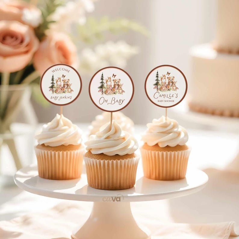 K&ouml;nnte beinhalten: Cupcakes mit wei&szlig;em Zuckerguss und dekorativen Toppern. Die Topper zeigen Illustrationen von Waldtieren und den Text &bdquo;Welcome Baby Jenkins&ldquo;, &bdquo;Oh Baby&ldquo; und &bdquo;Camille's Baby Shower&ldquo;. Die Cupcakes stehen auf einer wei&szlig;en Tortenplatte.