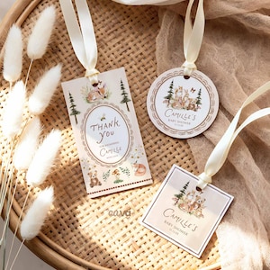 K&ouml;nnte beinhalten: Drei Babyparty-Geschenkanh&auml;nger mit cremefarbenen B&auml;ndern. Die Anh&auml;nger zeigen Illustrationen von Waldtieren und den Text "Thank You" und "Camille's Baby Shower". Die Anh&auml;nger liegen auf einem geflochtenen Tablett.