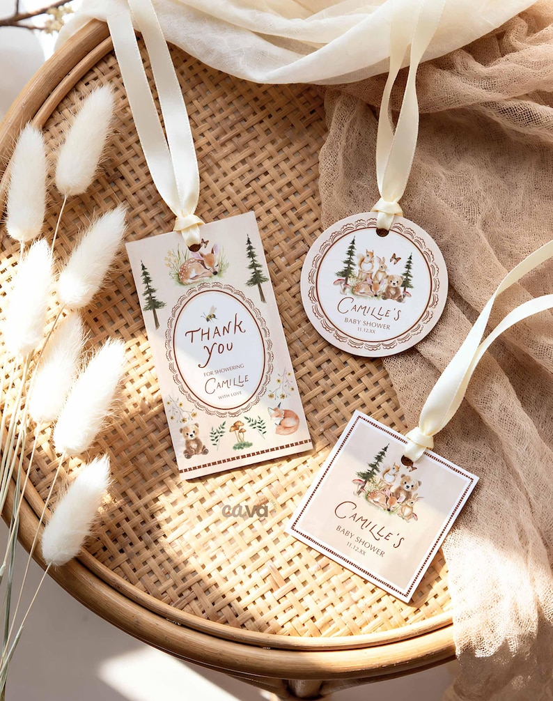 K&ouml;nnte beinhalten: Drei Babyparty-Geschenkanh&auml;nger mit cremefarbenen B&auml;ndern. Die Anh&auml;nger zeigen Illustrationen von Waldtieren und den Text "Thank You" und "Camille's Baby Shower". Die Anh&auml;nger liegen auf einem geflochtenen Tablett.