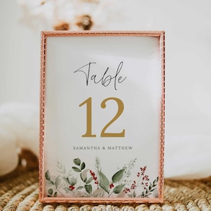 May include: A gold framed table number card with a white background and a gold number 12. The text "Table" is written in black cursive script above the number. The text "Samantha & Matthew" is written in black below the number. The card has a floral design with green leaves and red berries.