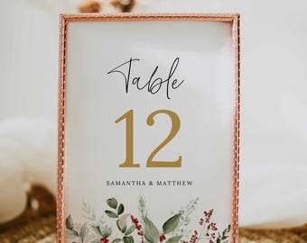 Pode incluir: Um cartão de número de mesa com moldura dourada, fundo branco e um número 12 dourado. O texto "Table" está escrito em caligrafia preta acima do número. O texto "Samantha & Matthew" está escrito em preto abaixo do número. O cartão tem um design floral com folhas verdes e bagas vermelhas.