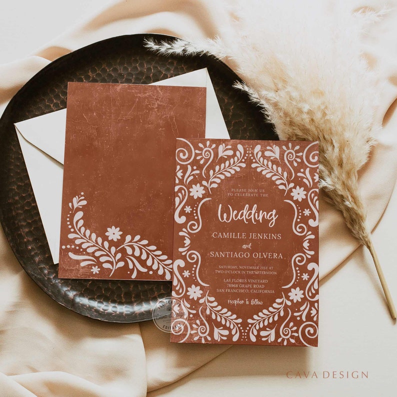 May include: Terracotta-colored wedding invitation suite with white floral design. The invitation features the word "Wedding" and the names Camille Jenkins and Santiago Olvera. Includes an envelope and a matching card.