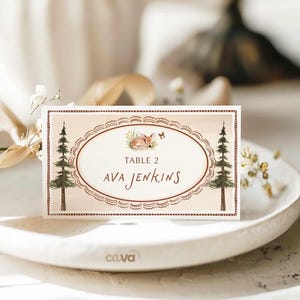 K&ouml;nnte beinhalten: Eine rechteckige Tischkarte mit Waldthema. Die Karte zeigt ein ovales Design mit einer Illustration eines schlafenden Fuchses, zwei Kiefern und dem Text "TABLE 2 AVA JENKINS" in brauner Schrift. Die Karte hat einen dekorativen Rand.