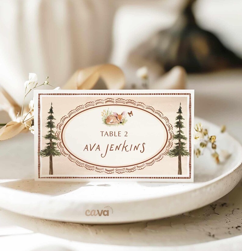K&ouml;nnte beinhalten: Eine rechteckige Tischkarte mit Waldthema. Die Karte zeigt ein ovales Design mit einer Illustration eines schlafenden Fuchses, zwei Kiefern und dem Text "TABLE 2 AVA JENKINS" in brauner Schrift. Die Karte hat einen dekorativen Rand.