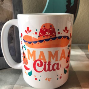 May include: White ceramic mug with a colorful design featuring a sombrero, guitars, and the text "Mama Cita".