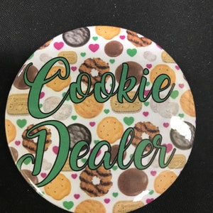 May include: A round button with a white background and a green design. The button features a variety of Girl Scout cookie images and the words "Cookie Dealer" in green cursive.