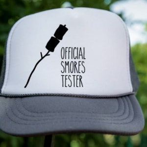 May include: A white and gray trucker hat with a black silhouette of a marshmallow on a stick and the text "OFFICIAL SMORES TESTER".