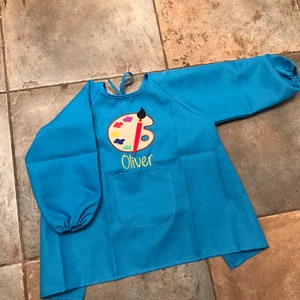 May include: A blue long-sleeved smock with a paint palette and paintbrush design. The smock has a pocket and the name "Oliver" embroidered on the front.