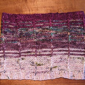 Puede incluir: Un cuello tejido a mano en tonos morado, rosa y crema. El cuello presenta un tejido de canalé vertical y un efecto degradado, pasando de un morado oscuro en la parte superior a un crema más claro en la parte inferior. El hilo tiene sutiles motas de verde azulado y verde.