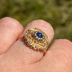 May include: A gold ring with an elaborate, filigree pattern. The ring has a central, round, dark blue gemstone. The ring is set against a blurred green background.