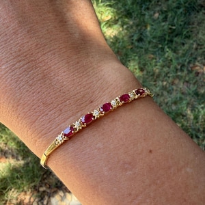 Puede incluir: Una pulsera de oro con una serie de rubíes rojos de corte ovalado que se alternan con pequeñas piedras de acento redondas y transparentes. La pulsera tiene un diseño articulado y un acabado en oro pulido, creando una línea continua.