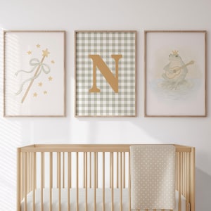 May include: A nursery scene featuring a light wood crib with a beige polka dot blanket. Above the crib are three framed prints: a magic wand, a letter "N" on a gingham background, and a frog playing a lute. The room has a neutral color palette.