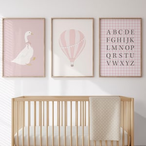 May include: A nursery scene featuring a light wood crib with a white mattress and a beige blanket with white polka dots. Above the crib are three framed prints: a pink goose, a pink hot air balloon, and a pink alphabet chart.