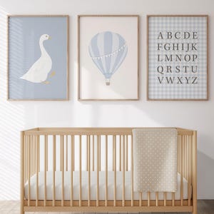 May include: A nursery scene featuring a light wood crib with a white mattress and a beige blanket with white polka dots. Above the crib are three framed prints: a goose, a hot air balloon, and an alphabet chart. The prints have a soft, pastel color palette.